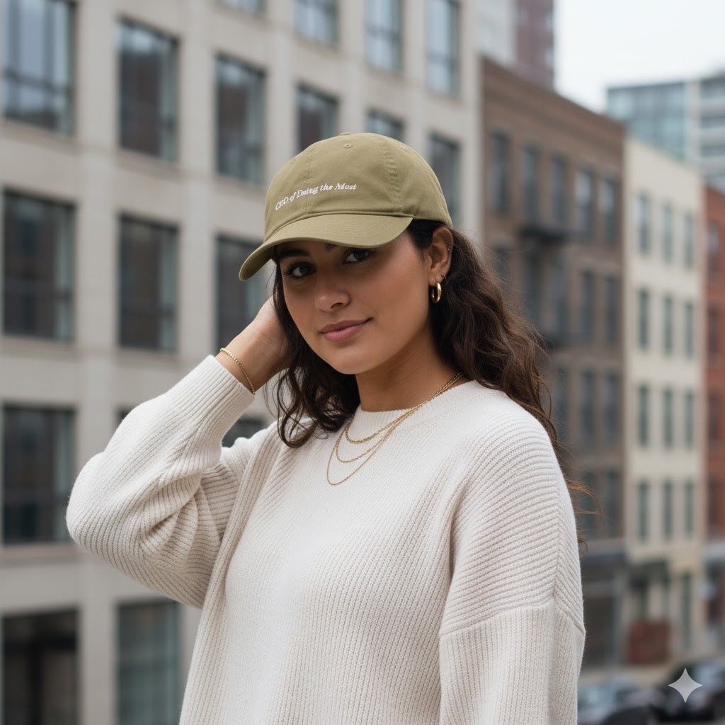 Woman wearing an olive cap that says "ceo of doing the most" and sweater in an urban setting