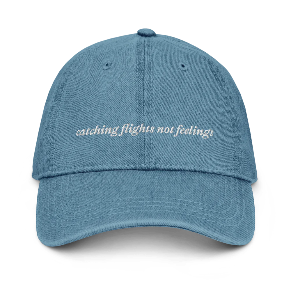 denim baseball hat with 'catching flights not feelings' text embroidered in white on a white background