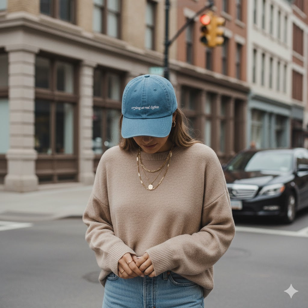 Person wearing a denim baseball hat that says "crying at redlights" and beige sweater on a city street