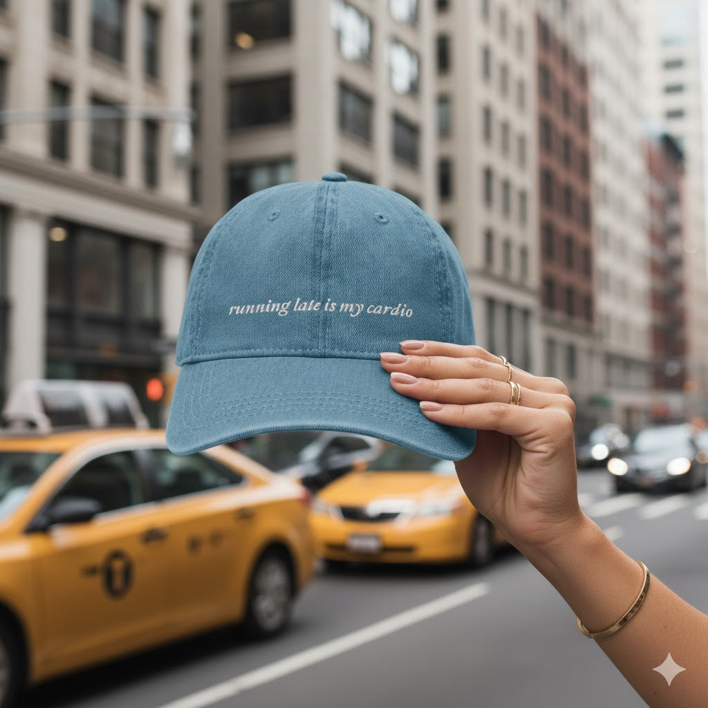 denim cap with text saying "running late is my cardio" held up in front of a city street with yellow taxis.