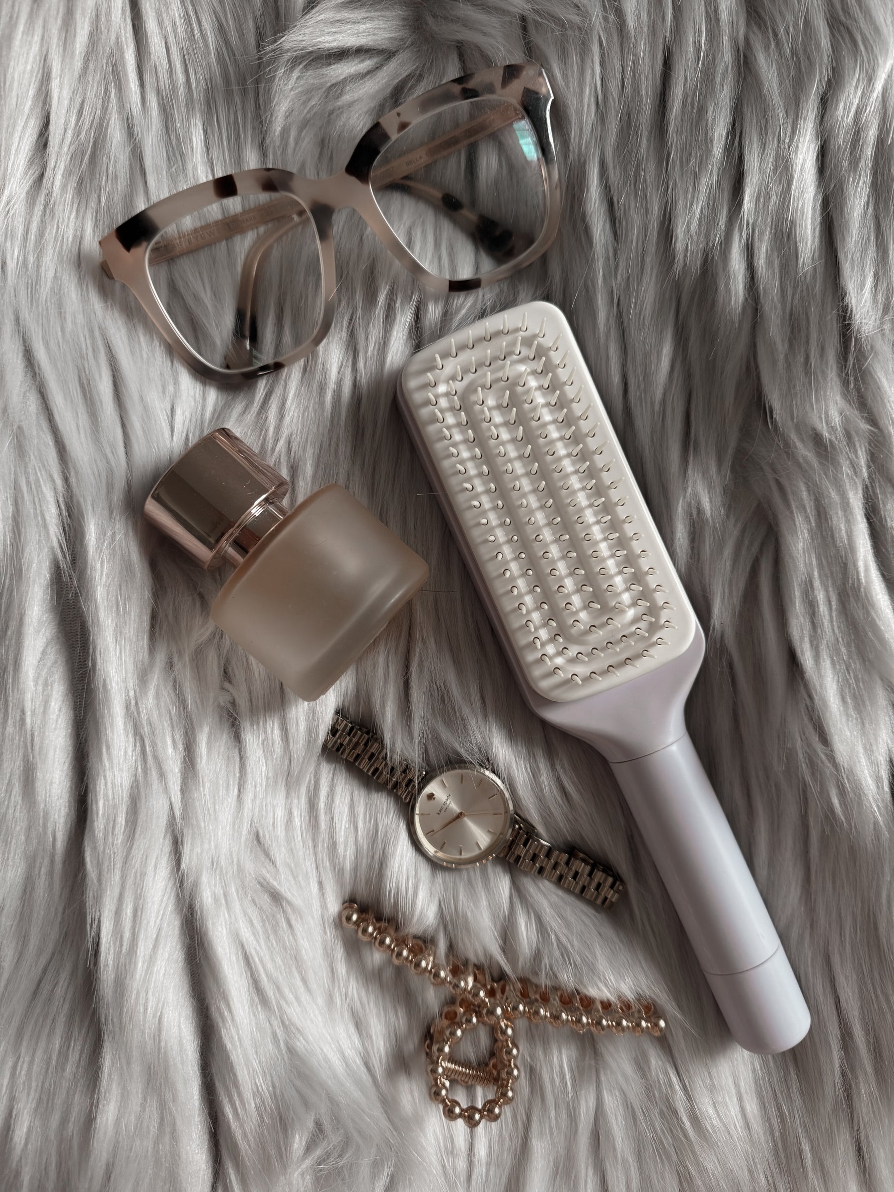 purple self cleaning hairbrush, sunglasses, bottle, watch, and bracelet on a fur surface
