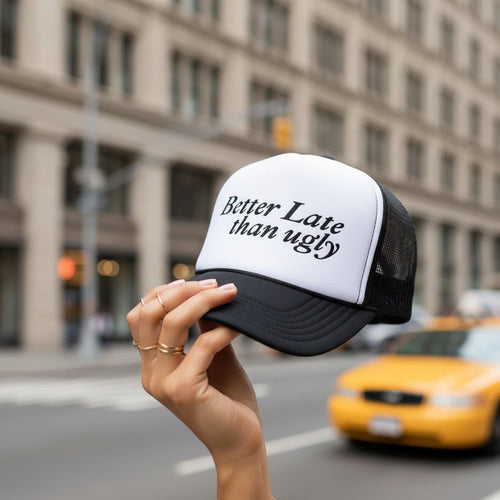 Hand holding a cap with 'Better Late than Ugly' text in an urban setting with a taxi in the background.