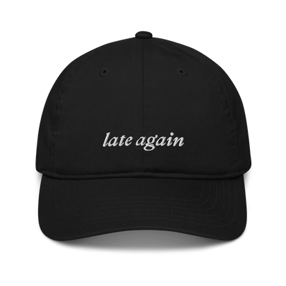 Black baseball hat with 'late again' text embroidered in white on a white background