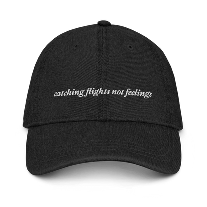 Black denim baseball hat with 'catching flights not feelings' text embroidered in white on a white background