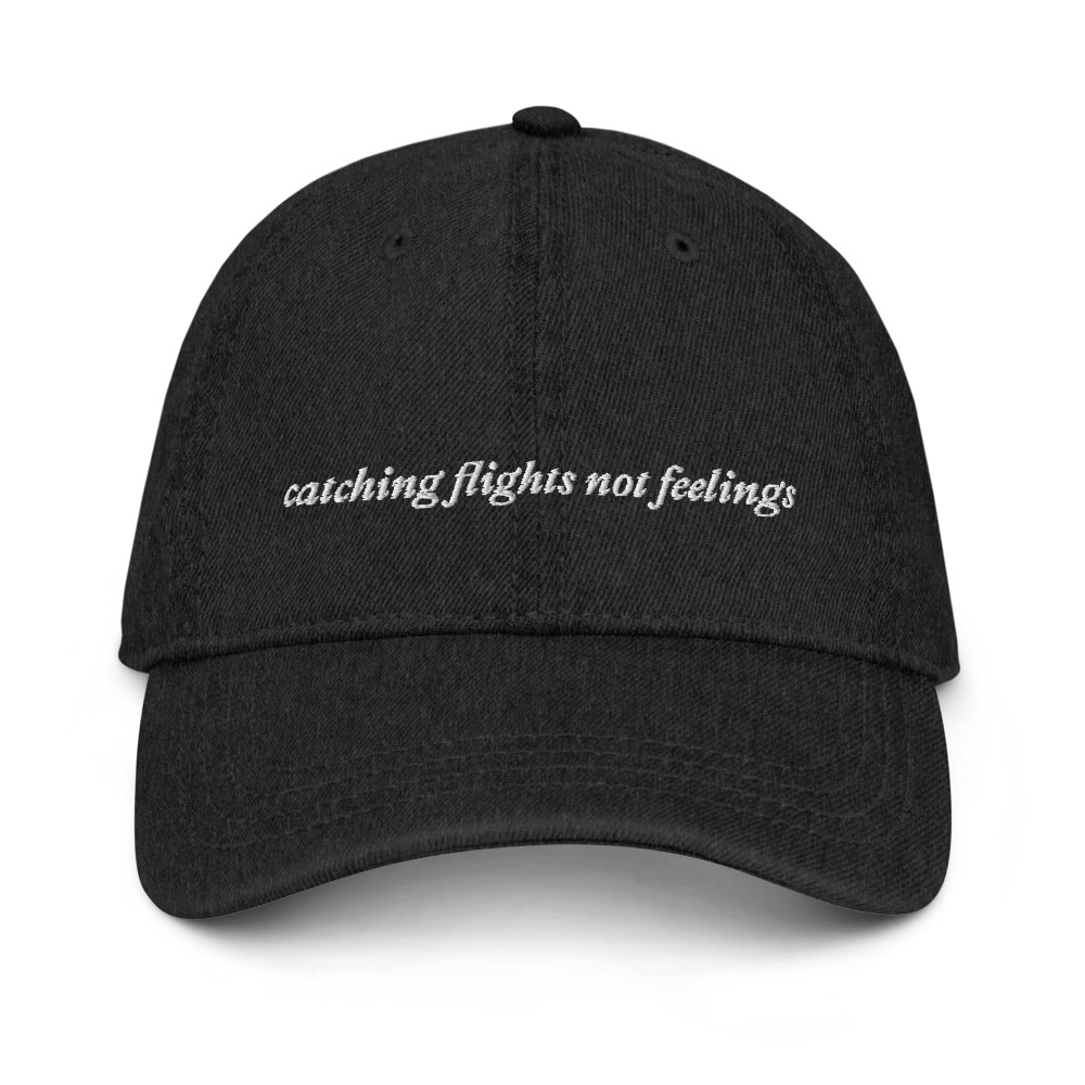 Black denim baseball hat with 'catching flights not feelings' text embroidered in white on a white background