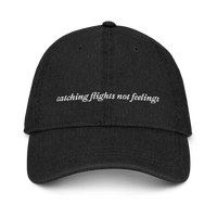 Black denim baseball hat with 'catching flights not feelings' text embroidered in white on a white background