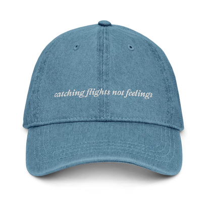  denim baseball hat with 'catching flights not feelings' text embroidered in white on a white background