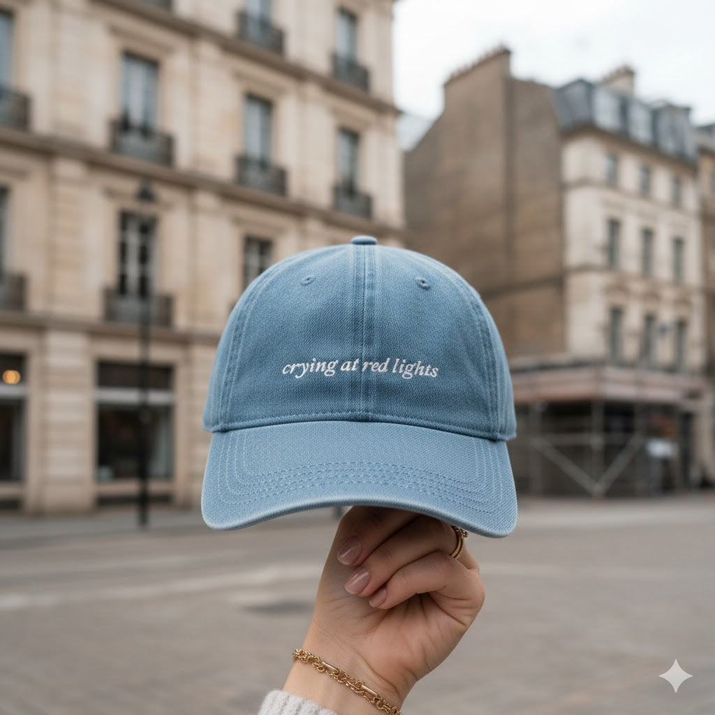 Light blue cap with 'crying at red lights' text held in front of a city street background
