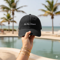 Black cap with 'Do Not Disturb' text held by a hand by a pool with palm trees in the background