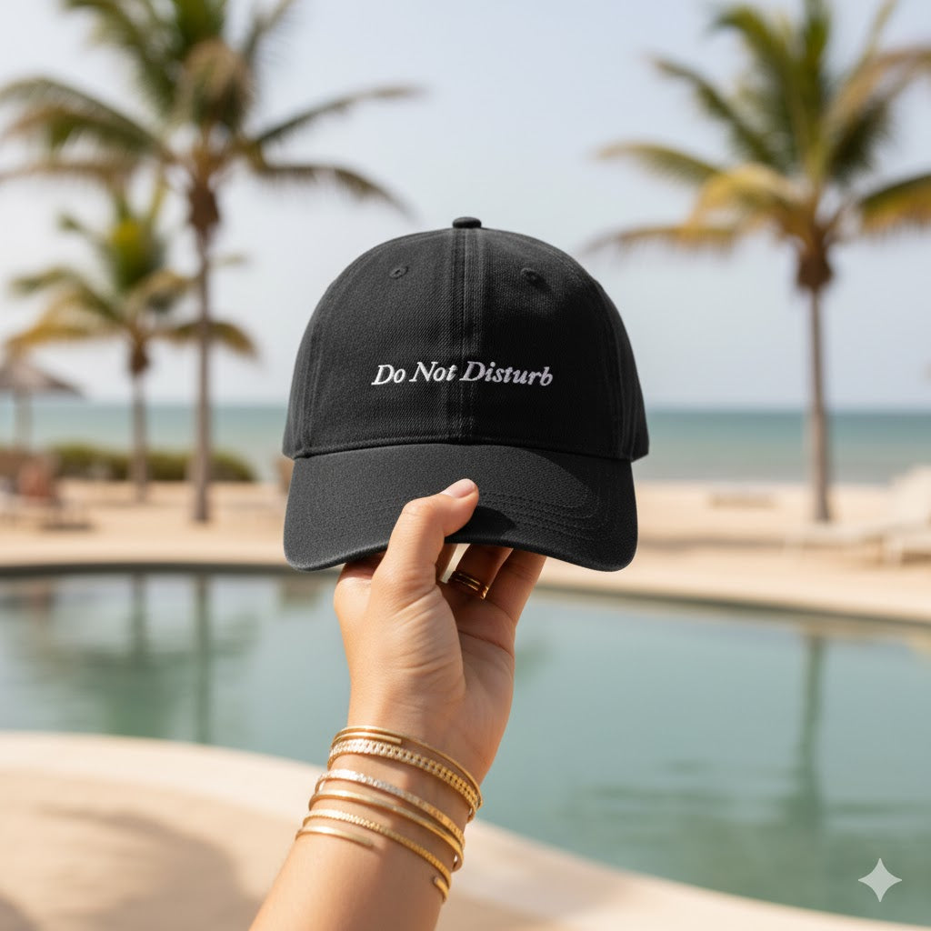 Black cap with 'Do Not Disturb' text held by a hand by a pool with palm trees in the background