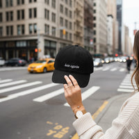 Person holding a black cap with 'late again' text on a city street.
