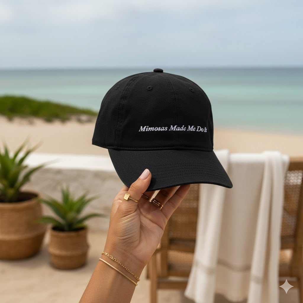 Black cap with 'Mimosas Made Me Do It' text held by a hand on a beach.