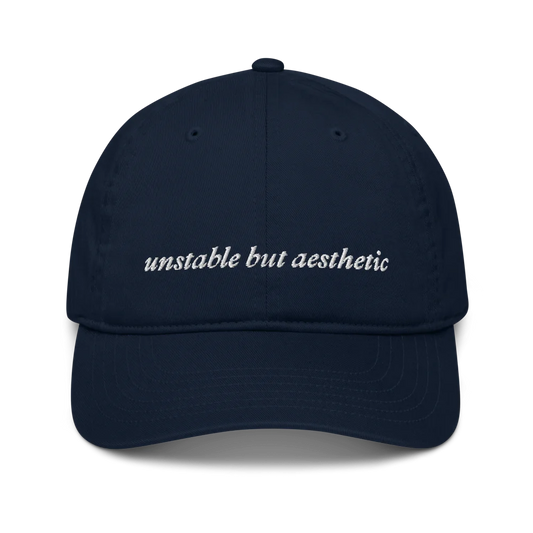 Navy blue baseball hat with 'unstable but aesthetic' text embroidered in white on a white background