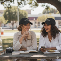 Two women sitting at a table outdoors, wearing matching black hats that say "this and not answering my texts".
