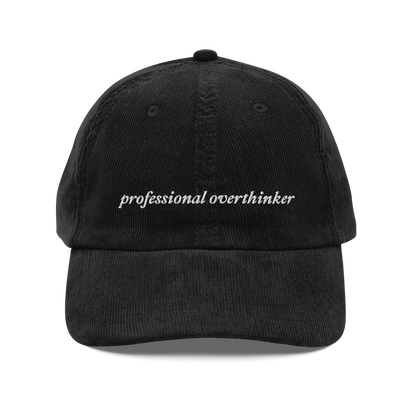 black corduroy baseball hat with 'professional overthinker' text embroidered in white on a white background