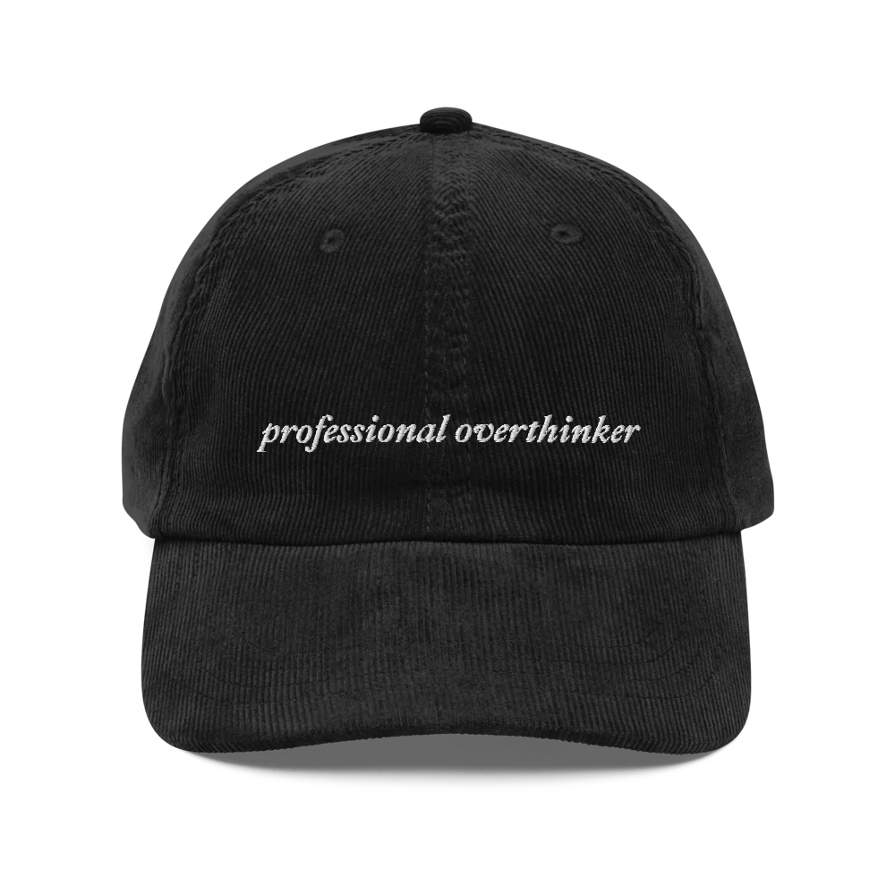 black corduroy baseball hat with 'professional overthinker' text embroidered in white on a white background