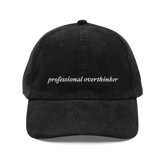 black corduroy baseball hat with 'professional overthinker' text embroidered in white on a white background