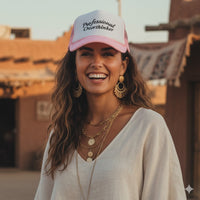 Woman wearing a pink and white trucker hat with text that says "professional overthinker" at a festival, surrounded by people and lights.