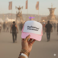 Hand holding a pink and white trucker hat with 'Professional Overthinker' text at a festival.