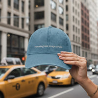 denim cap with text saying "running late is my cardio" held up in front of a city street with yellow taxis.