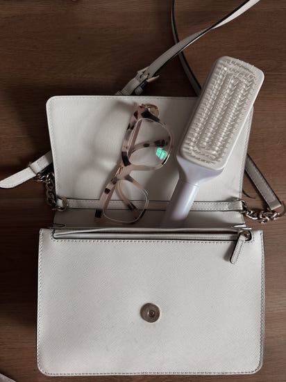 White handbag with glasses and a self cleaning hairbrush on a wooden surface