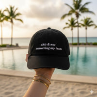 Black baseball hat with text that says "this and not answering my texts"  held by a person by a pool with palm trees in the background